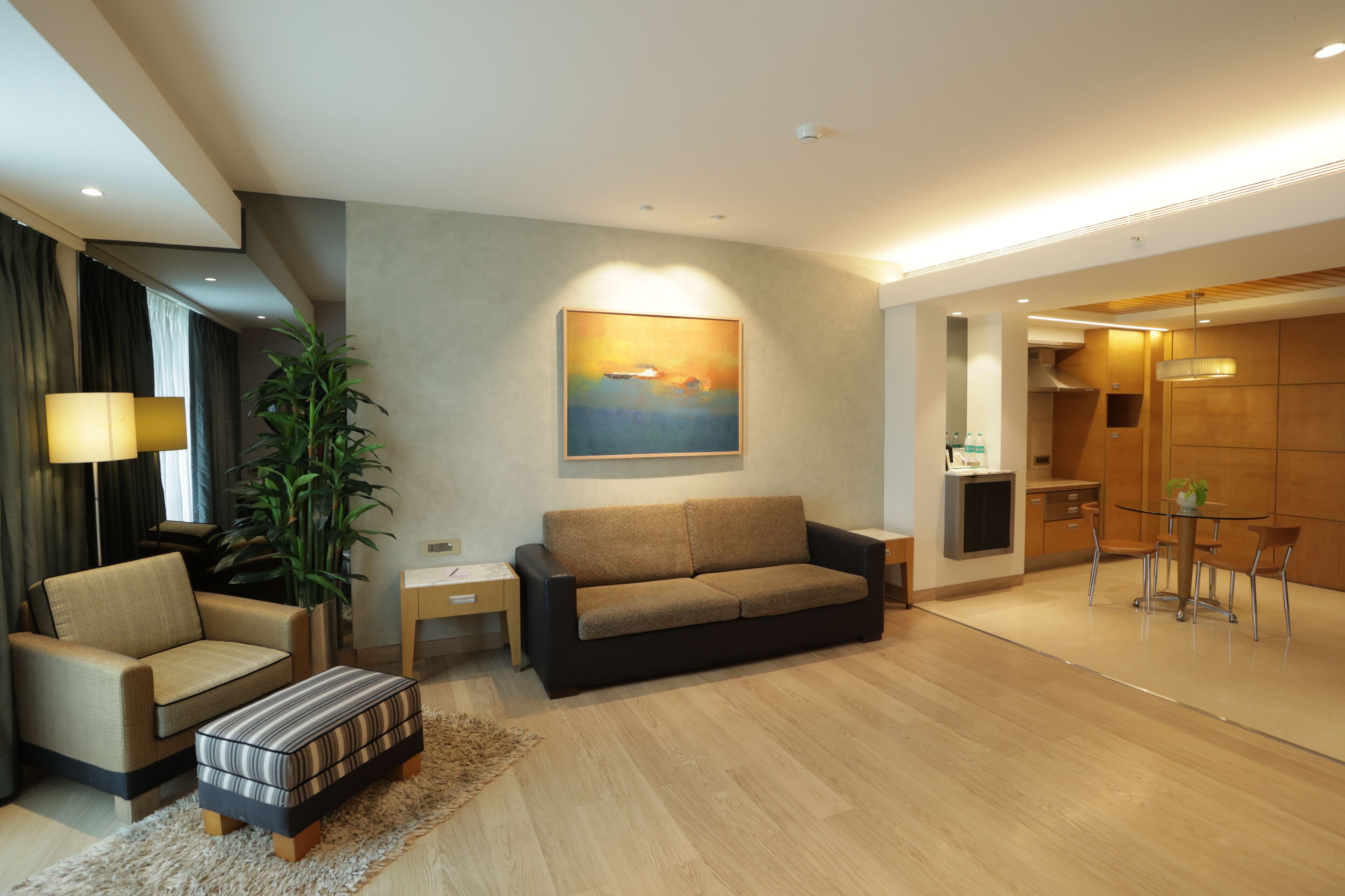 A modern living room with a sofa, armchair, and ottoman, a painting on the wall, and a dining area with a table and chairs.