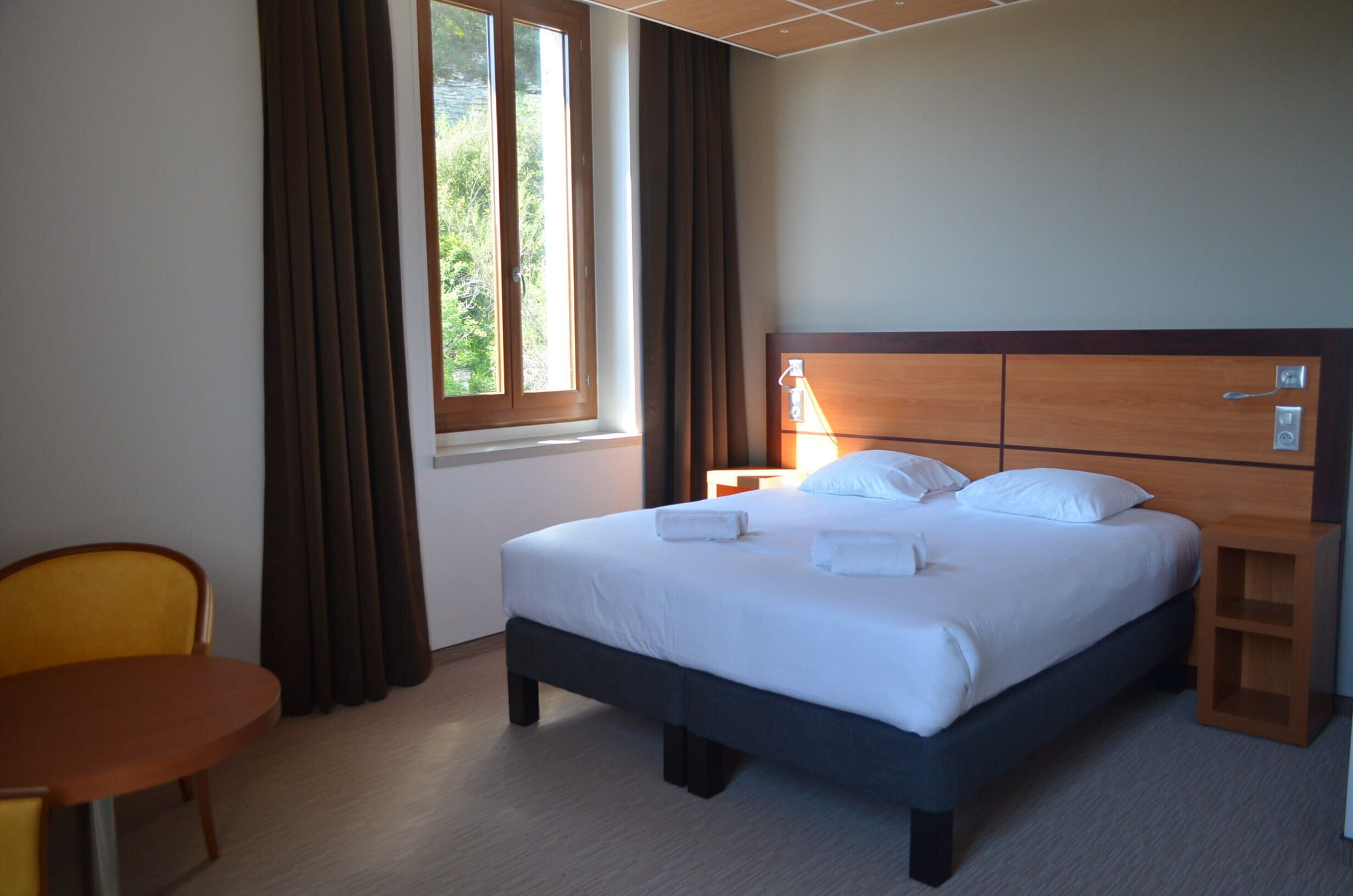 A hotel room with a bed, a bedside table, a chair, and a window with curtains.