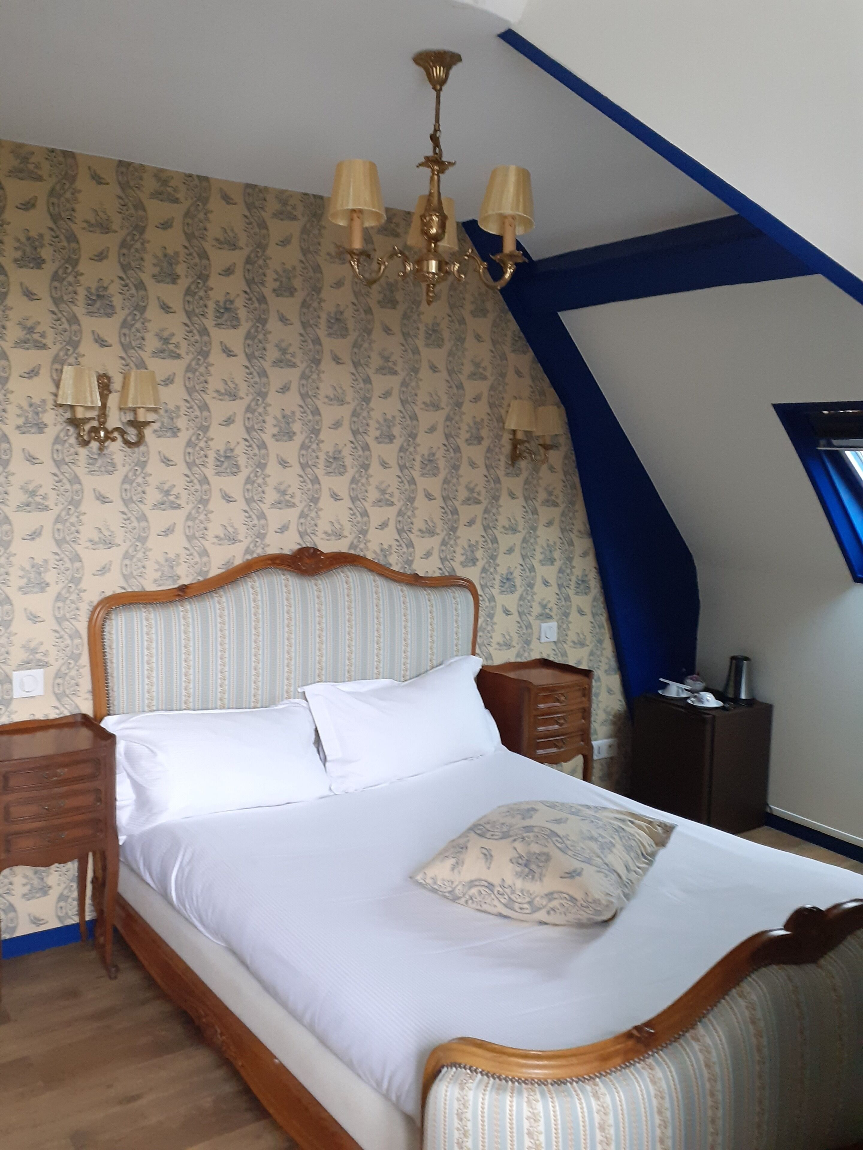 A bedroom with a wooden bed frame, white bedding, and a patterned pillow. There is a nightstand with a lamp, a dresser, and a window with a view.