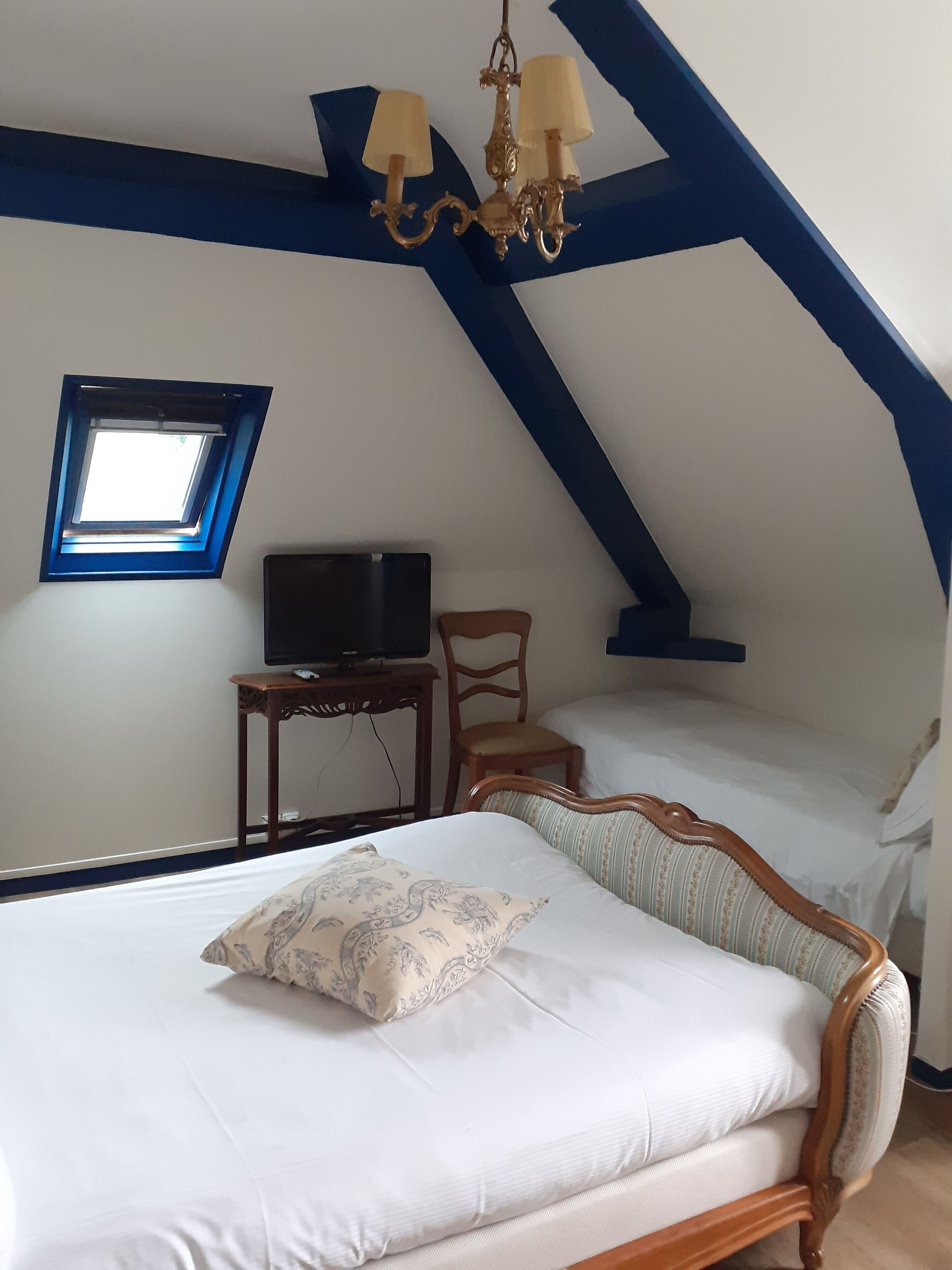 Une chambre mansardée avec un plafond en pente, un lit avec une literie blanche, une chaise en bois, une petite table avec un téléviseur et une fenêtre avec un cadre bleu.