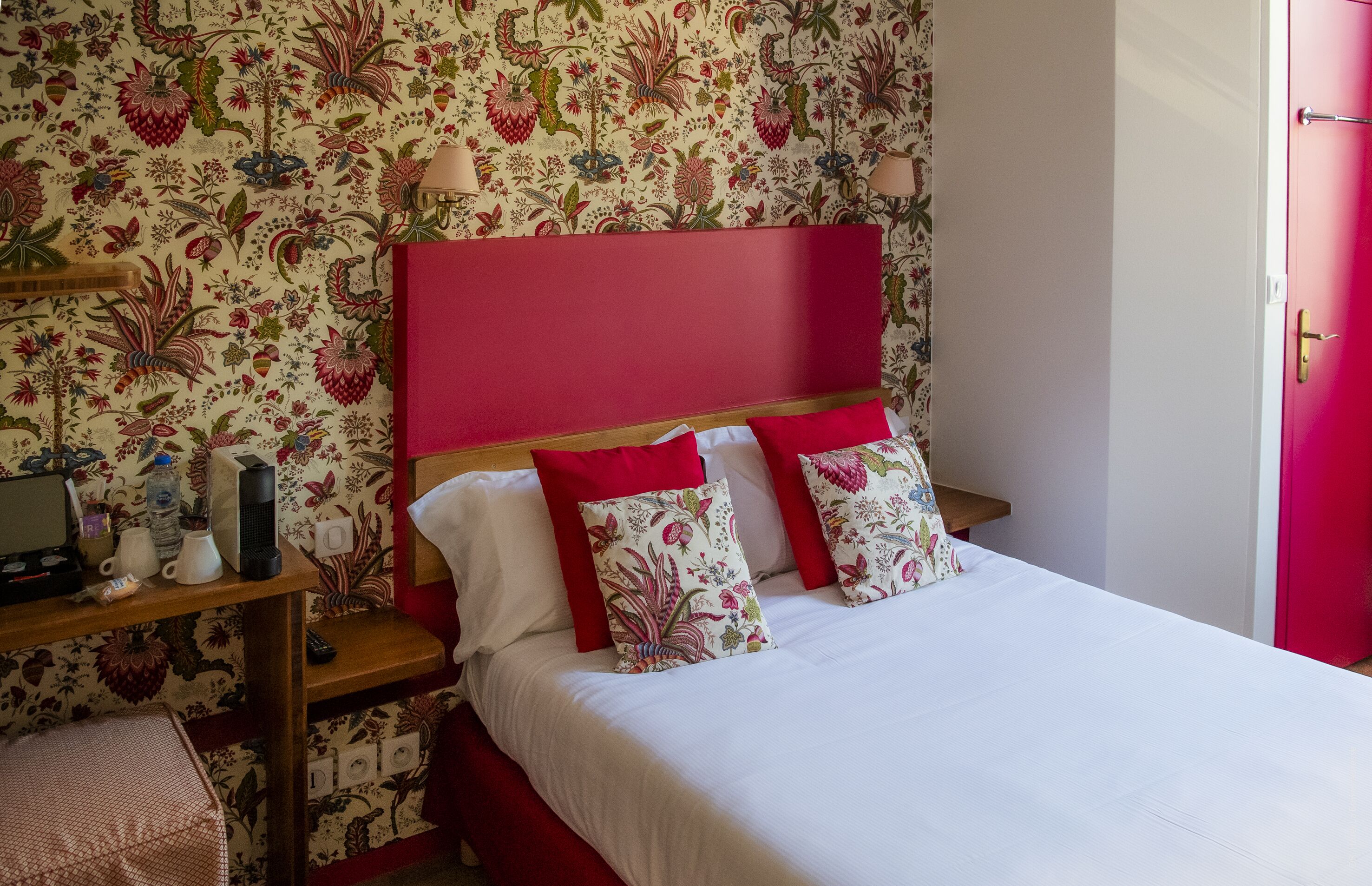 A neatly made bed with red and white bedding, a wooden nightstand with a lamp, and a floral wallpaper pattern.