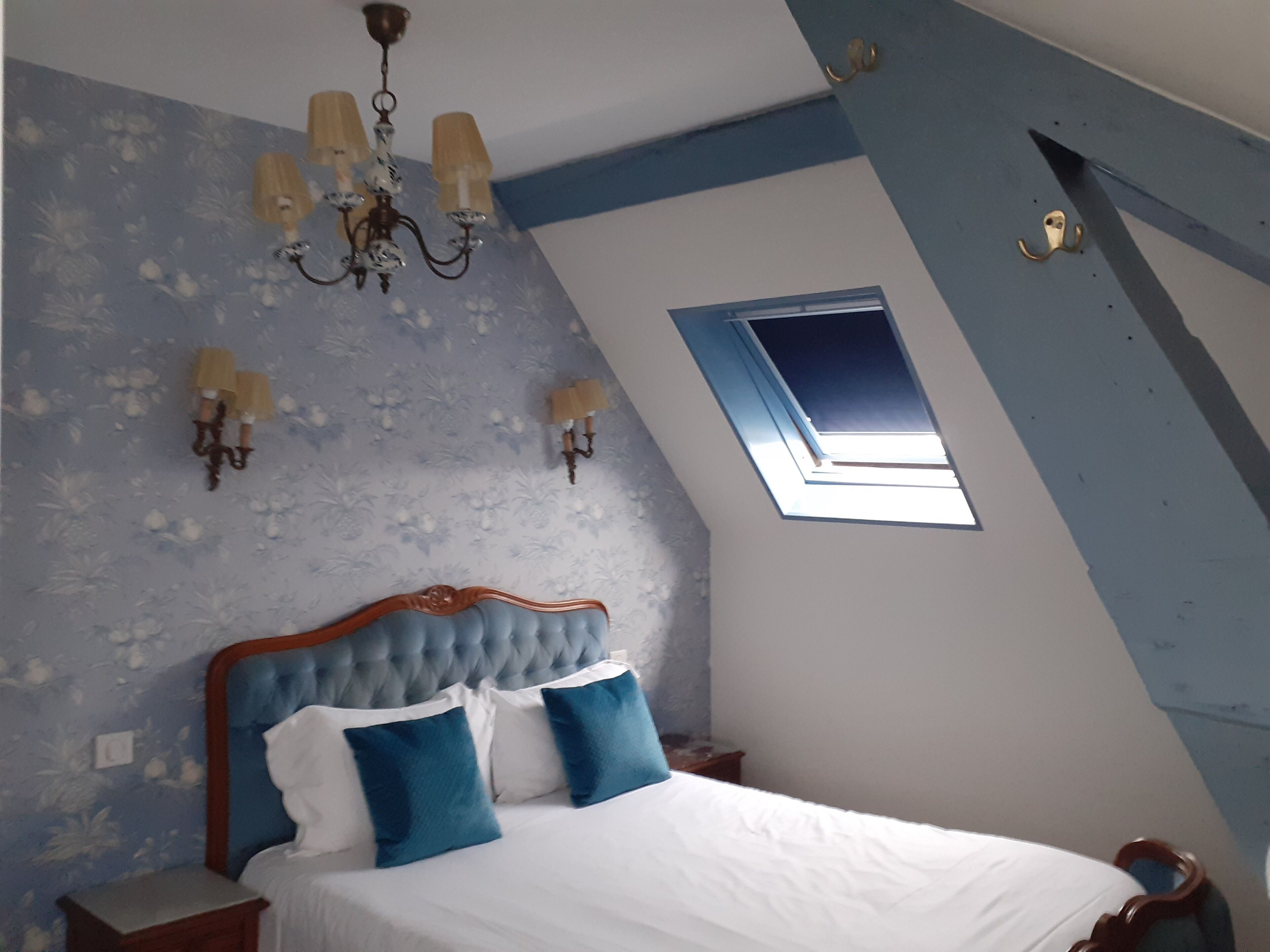 Ein Schlafzimmer mit schrägen Wänden, einem Oberlicht, einer Kristallleuchtre und einem Bett mit blau-weißer Bettwäsche.