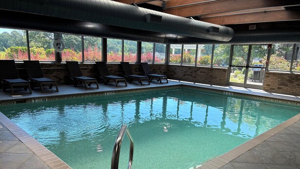 Una piscina interior con agua cristalina, una escalera metálica y sillas de descanso a la orilla.