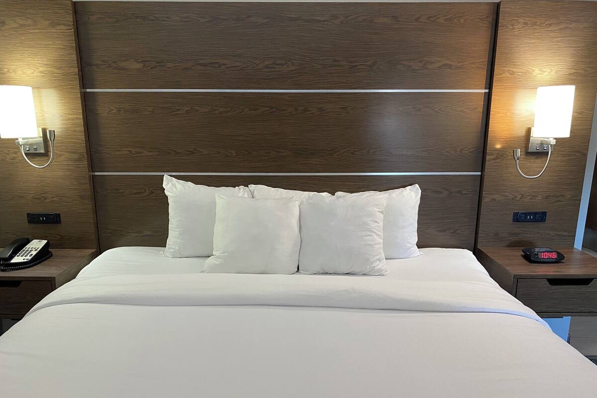 Een opgemaakt bed met witte lakens en kussens, omlijst door houten hoofdborden met wandlampen.