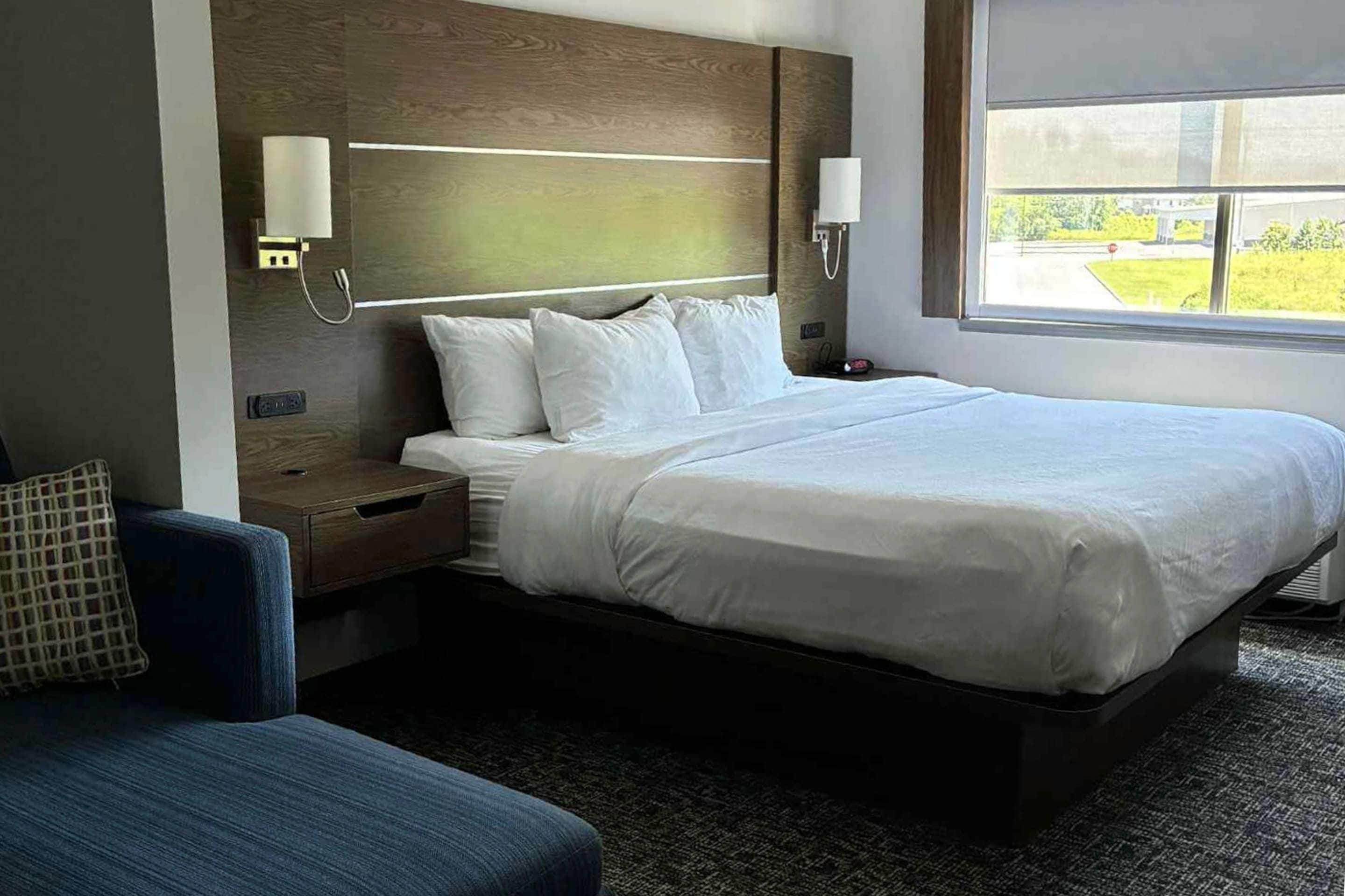Habitación de hotel con una cama grande, mesitas de noche, una silla y una ventana con vistas a zonas verdes.