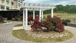 Un edificio con un espacio exterior techado que presenta una pérgola blanca, rodeado de arbustos florecientes rojos y un jardín ajardinado con una mezcla de césped y grava.