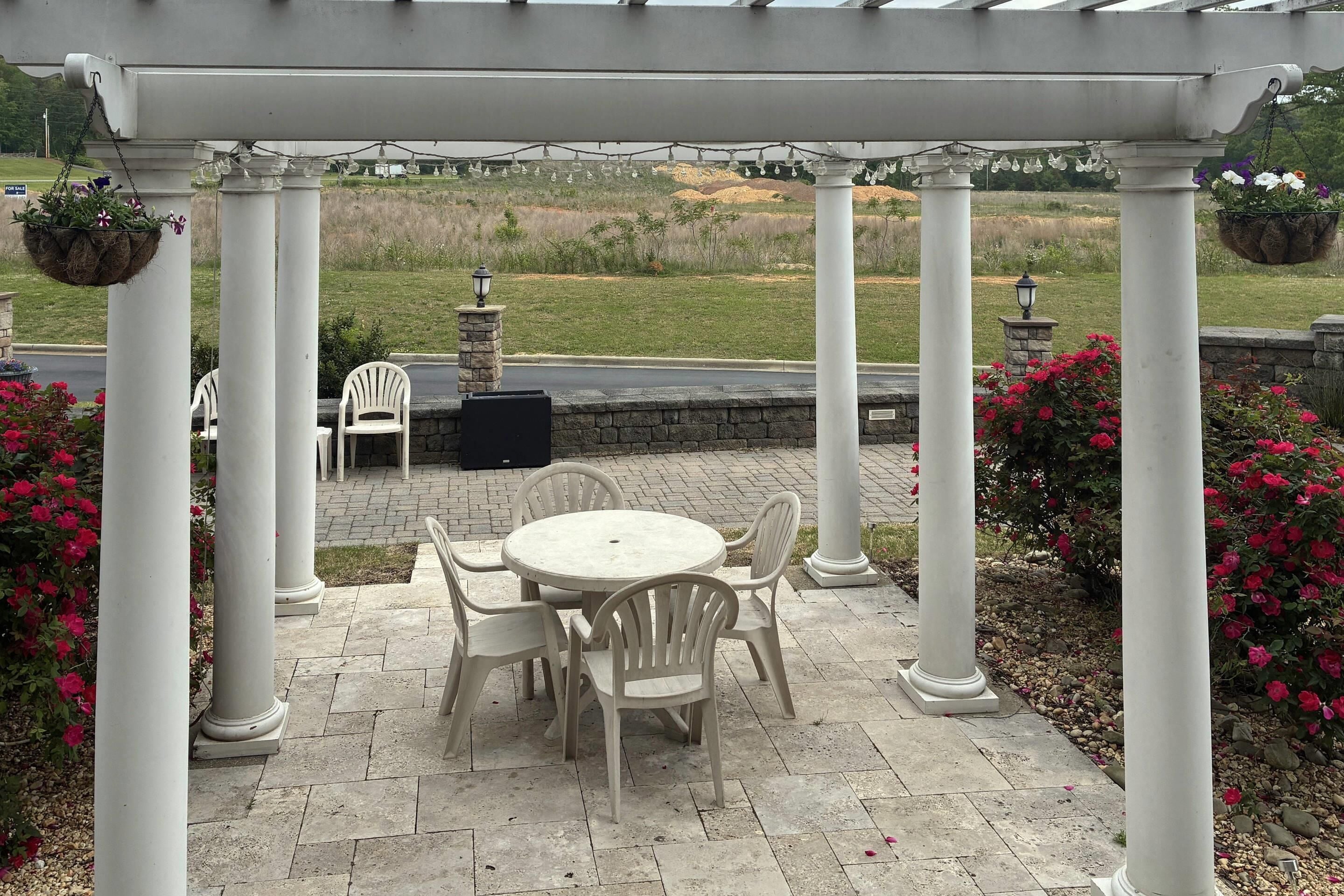 A covered patio area with a round table and chairs, white columns, hanging flower baskets, and a view of a grassy field.