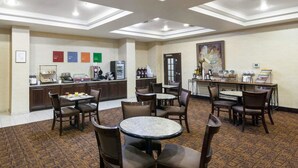 A hotel dining area with multiple tables and chairs, a kitchen counter with breakfast items, and a large painting on the wall. - Comfort Inn & Suites Regional Medical Center (Abilene)