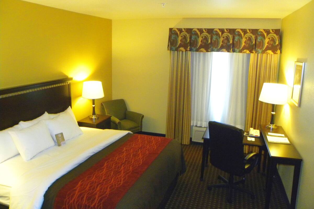 A hotel room with a bed, two bedside lamps, a desk with a chair, and a window with curtains.