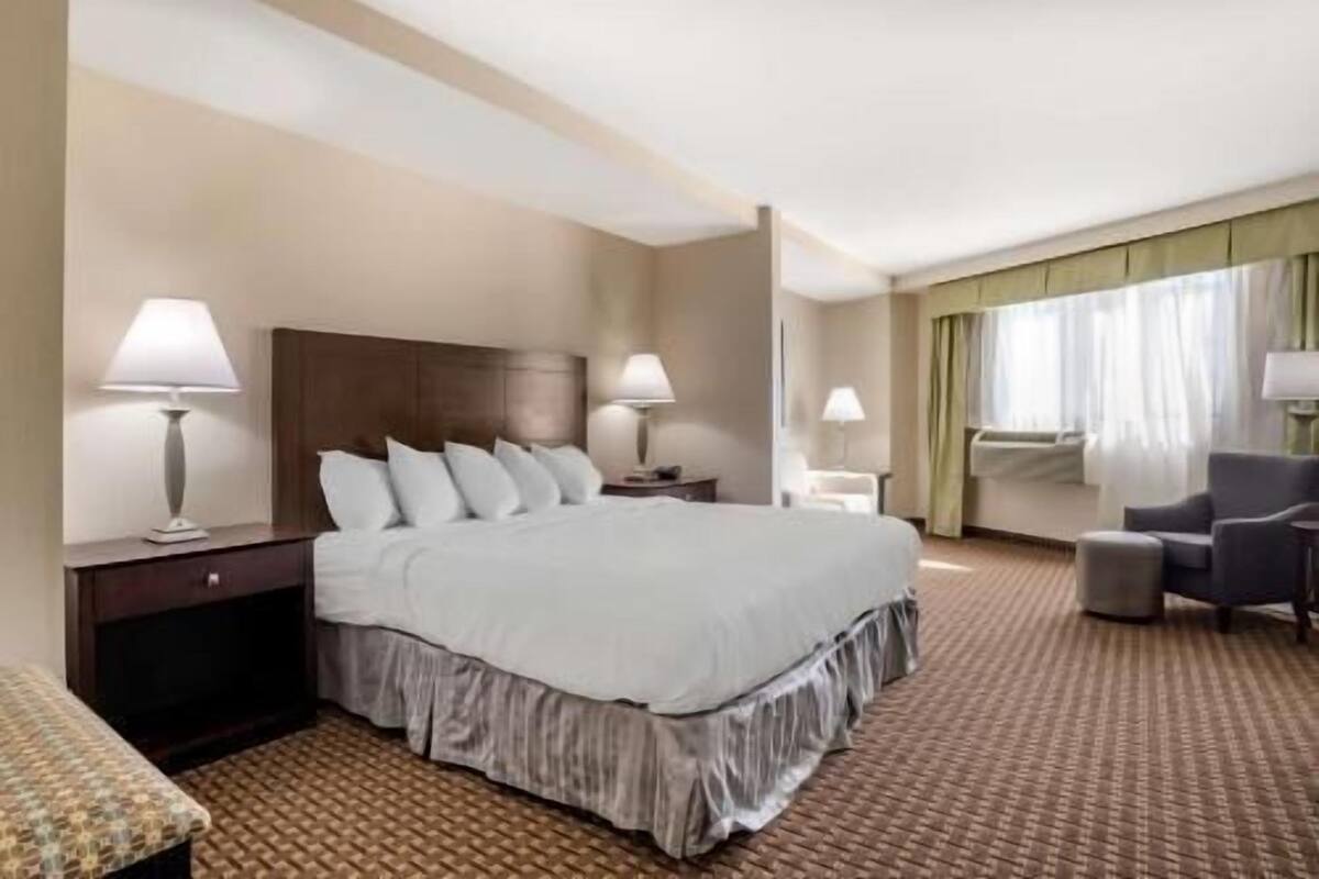 Een hotelkamer met een groot bed, twee nachtkastjes met lampen, een stoel, een klein tafeltje en een raam met gordijnen.