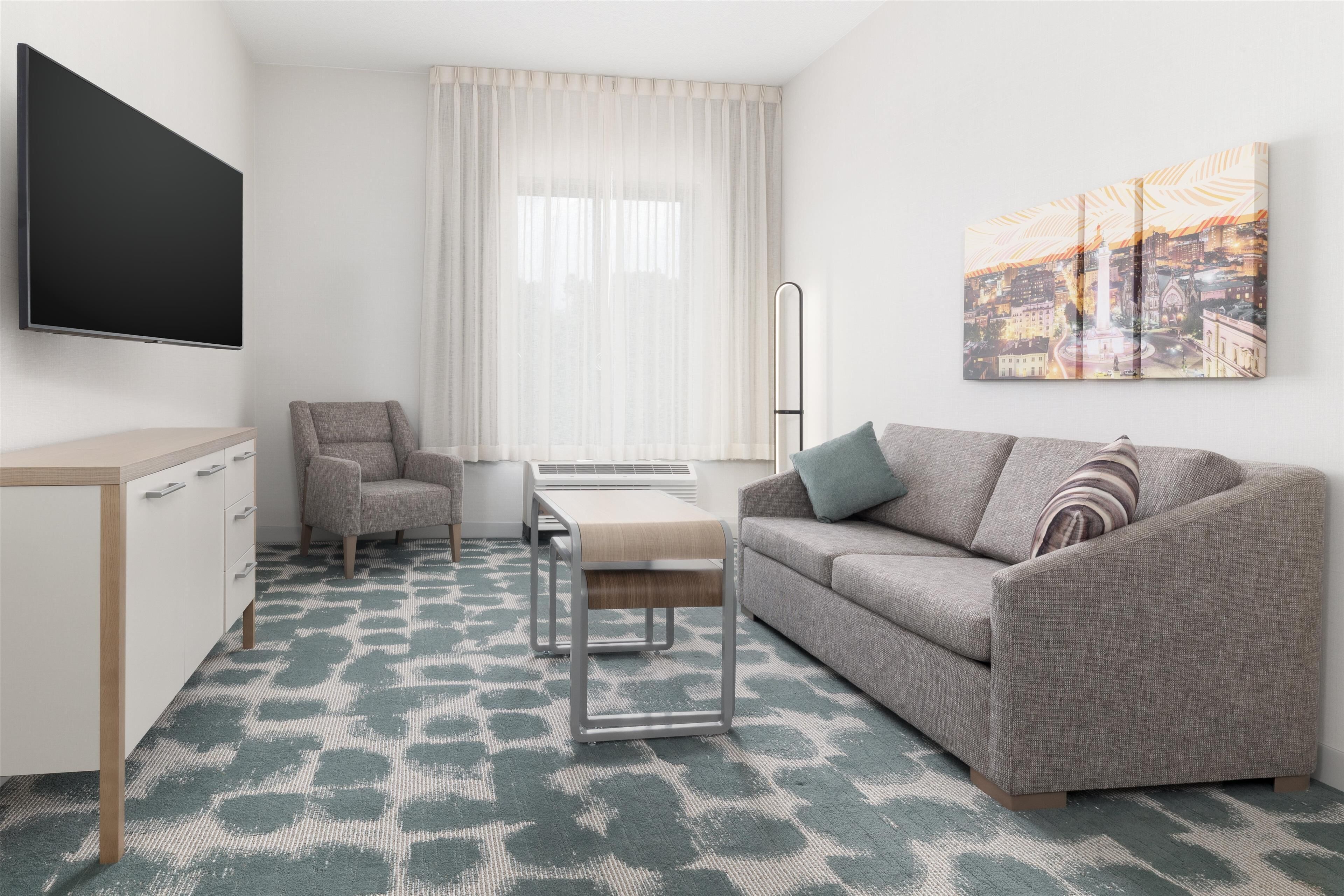Ein modernes Wohnzimmer mit einem Flachbildfernseher, einem grauen Sofa, einem grauen Sessel, einem kleinen Holztisch und einem Stadtbild an der Wand.