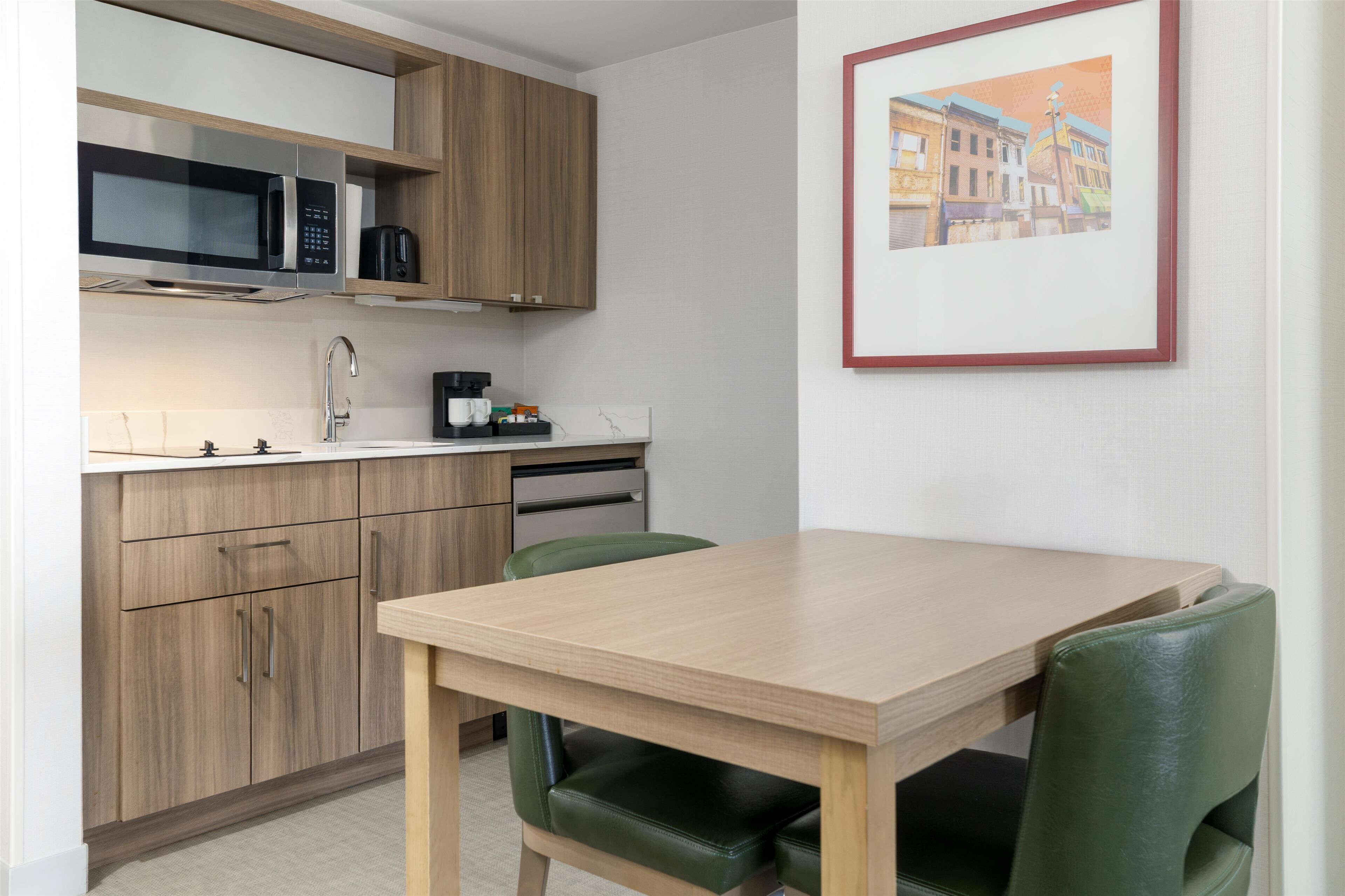 Een moderne keuken met houten kasten, een magnetron en een koffiezetapparaat. Er staat een eettafel met twee stoelen en er hangt een lijstje aan de muur.