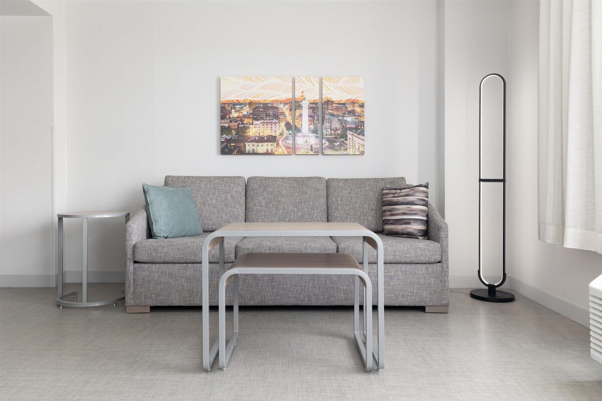 Un salon moderne avec un canapé gris, une petite table, un lampadaire et un tableau représentant un paysage urbain accroché au mur.