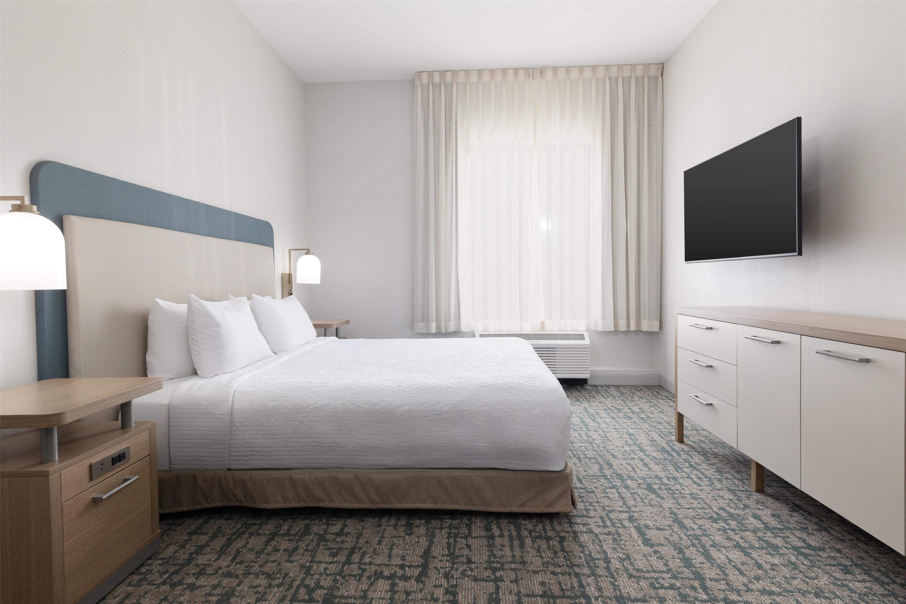 Una habitación de hotel con cama, mesitas de noche, televisión, escritorio y ventana con cortinas.