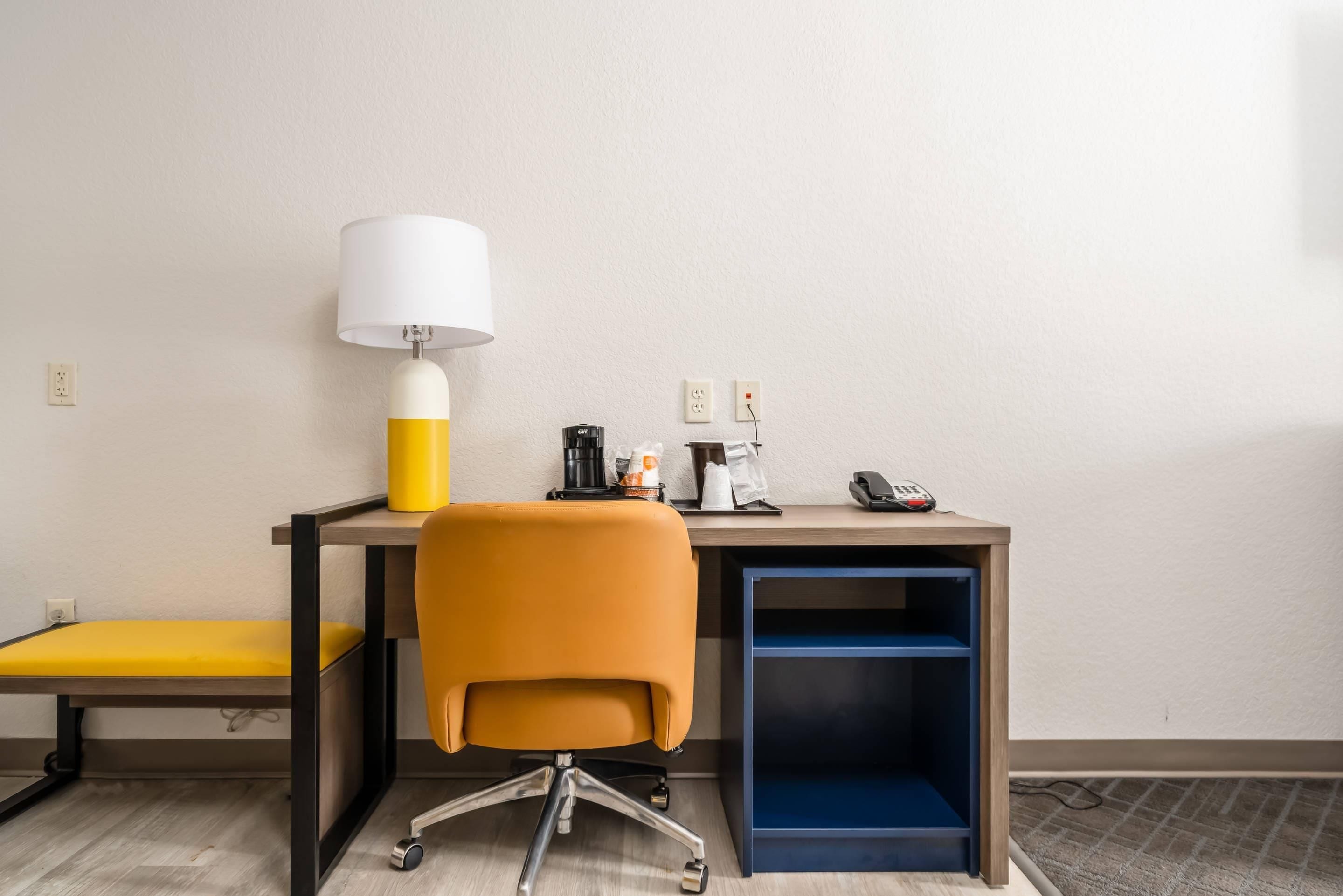 Habitación de hotel pequeña con escritorio, una silla amarilla, lámpara, cafetera y teléfono.