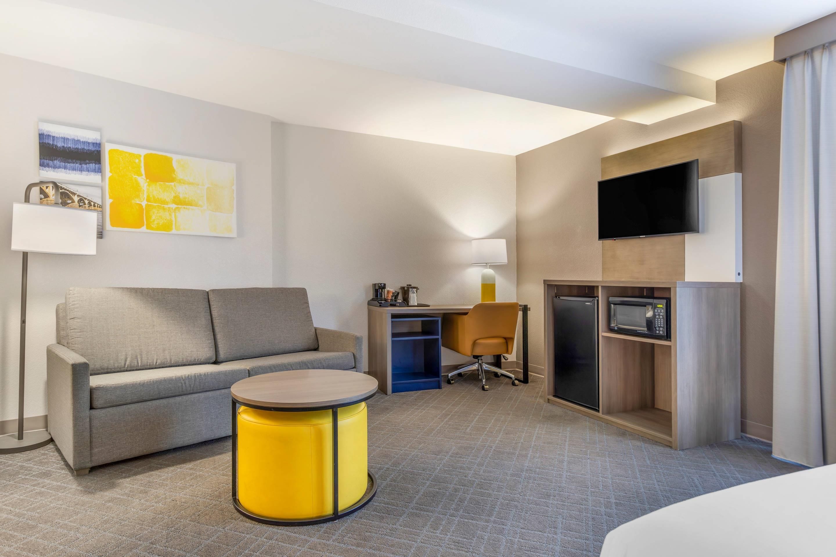 A hotel room with a sofa, a small round table, a desk with a chair, a television, and a microwave.