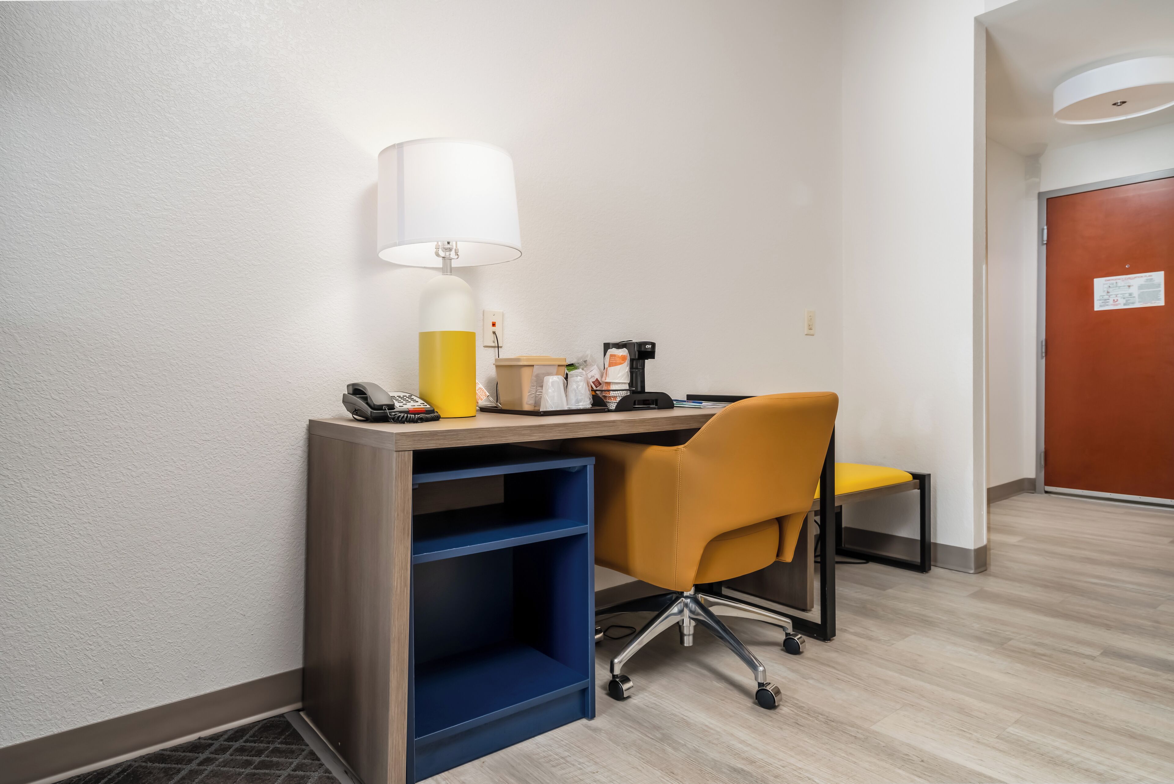 Habitación de hotel con escritorio, una silla amarilla, lámpara, teléfono y cafetera.