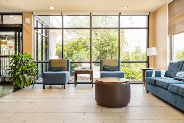 A modern lobby with large windows, a seating area with sofas and chairs, a round coffee table, and a potted plant.