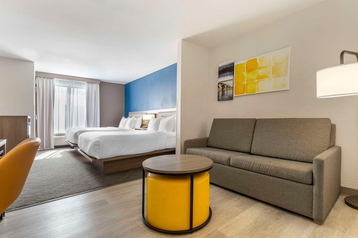 Une chambre d’hôtel avec un lit, un canapé, une table ronde jaune et une lampe.