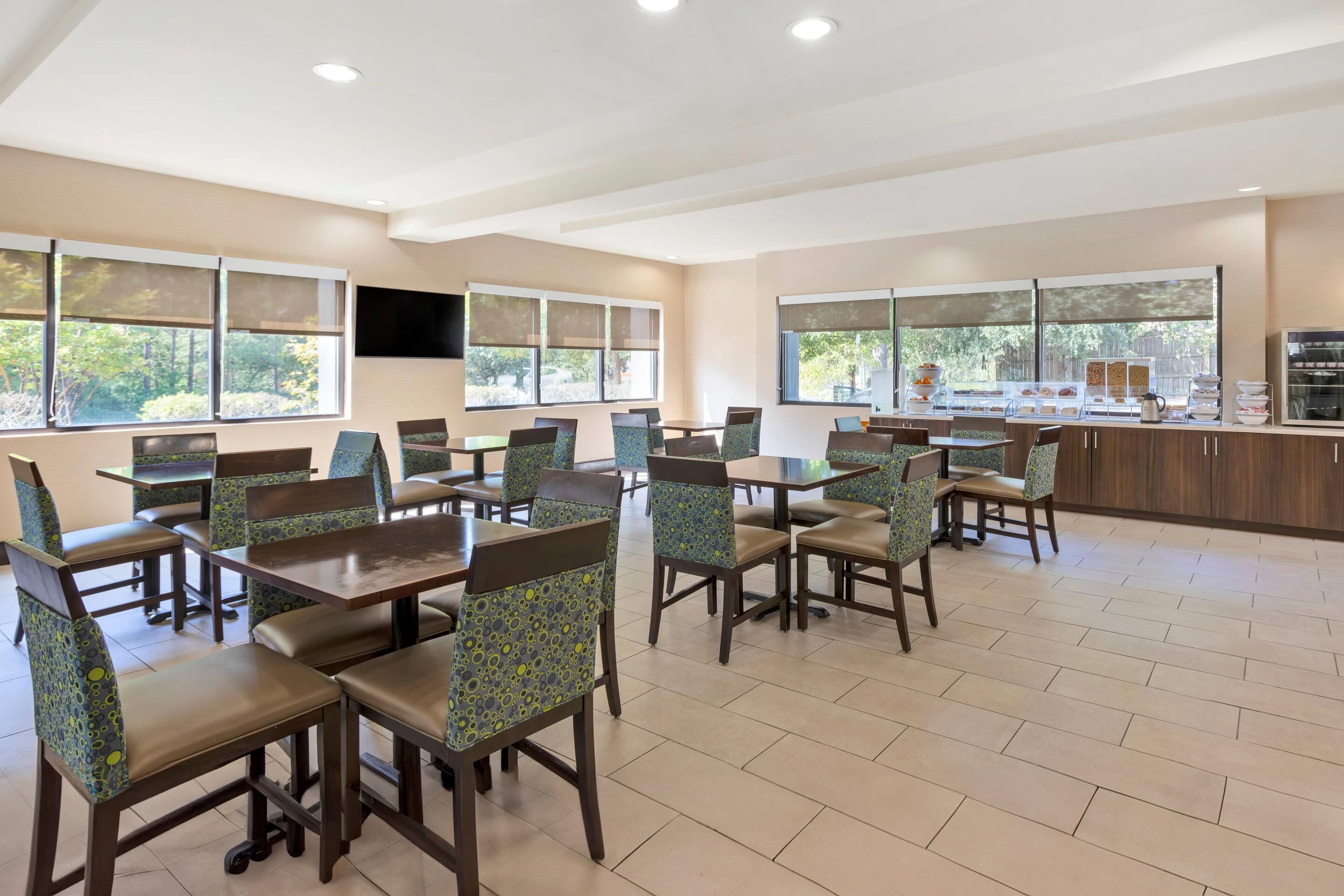 A spacious dining area with multiple tables and chairs, large windows with blinds, and a kitchen counter with various food items.