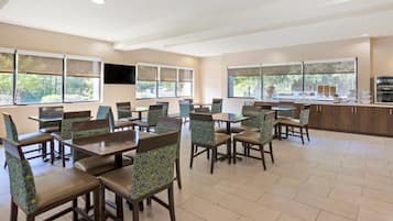 A spacious dining area with multiple tables and chairs, large windows with blinds, and a kitchen counter with various food items.