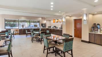 Uma área de refeições de hotel com várias mesas e cadeiras, um balcão com uma máquina de café e vista para o exterior através de grandes janelas.