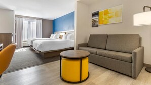Une chambre d’hôtel avec un lit, un canapé, un pouf jaune, une lampe et un tableau au mur.