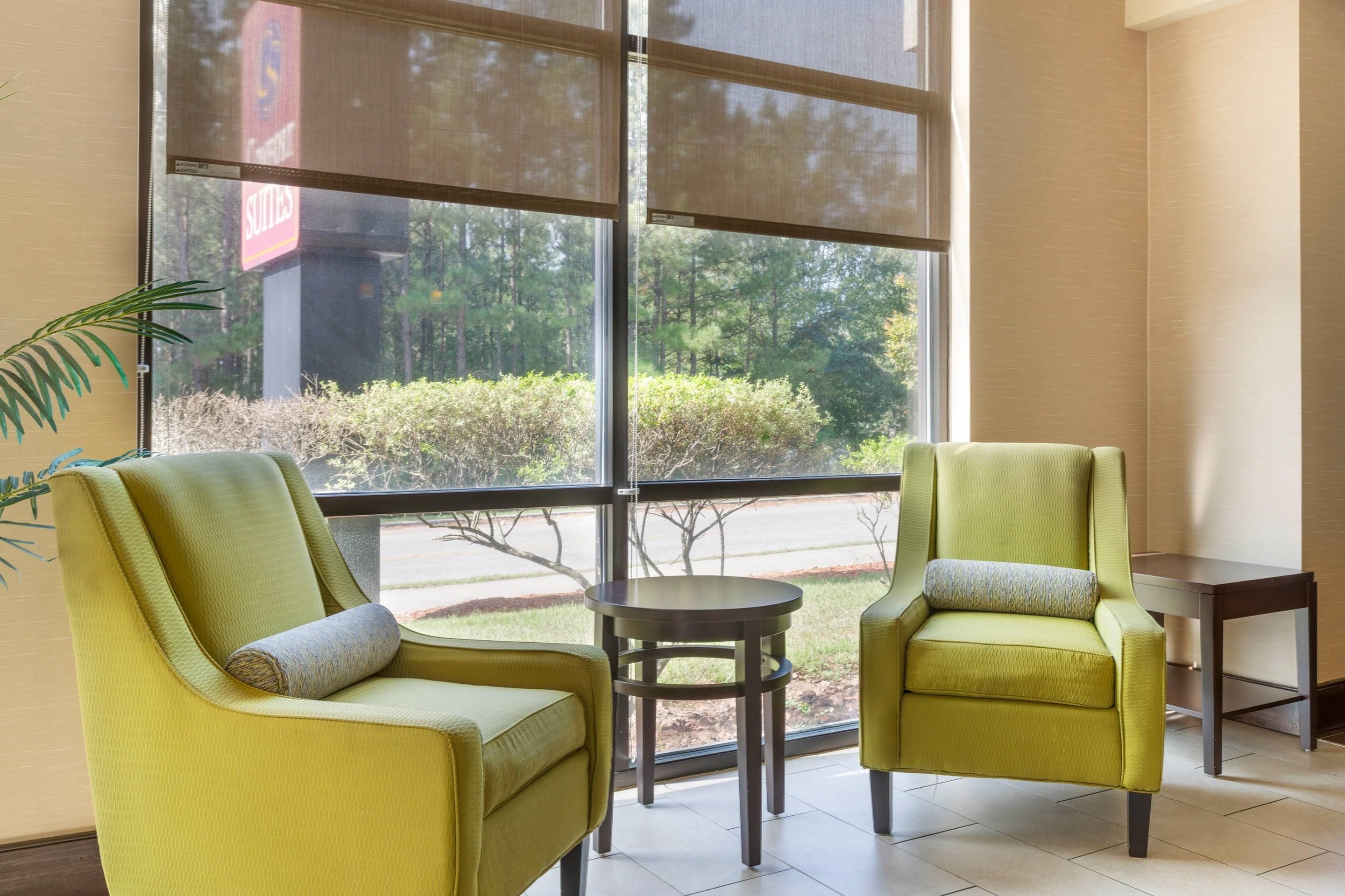 Une pièce avec deux fauteuils jaunes et une petite table, de grandes fenêtres donnant sur des arbres et une enseigne indiquant « Suits ».