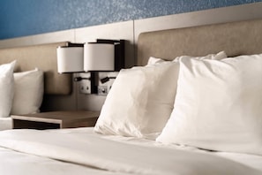 A neatly made bed with white linens and pillows, a wooden bedside table, and a wall-mounted lamp.