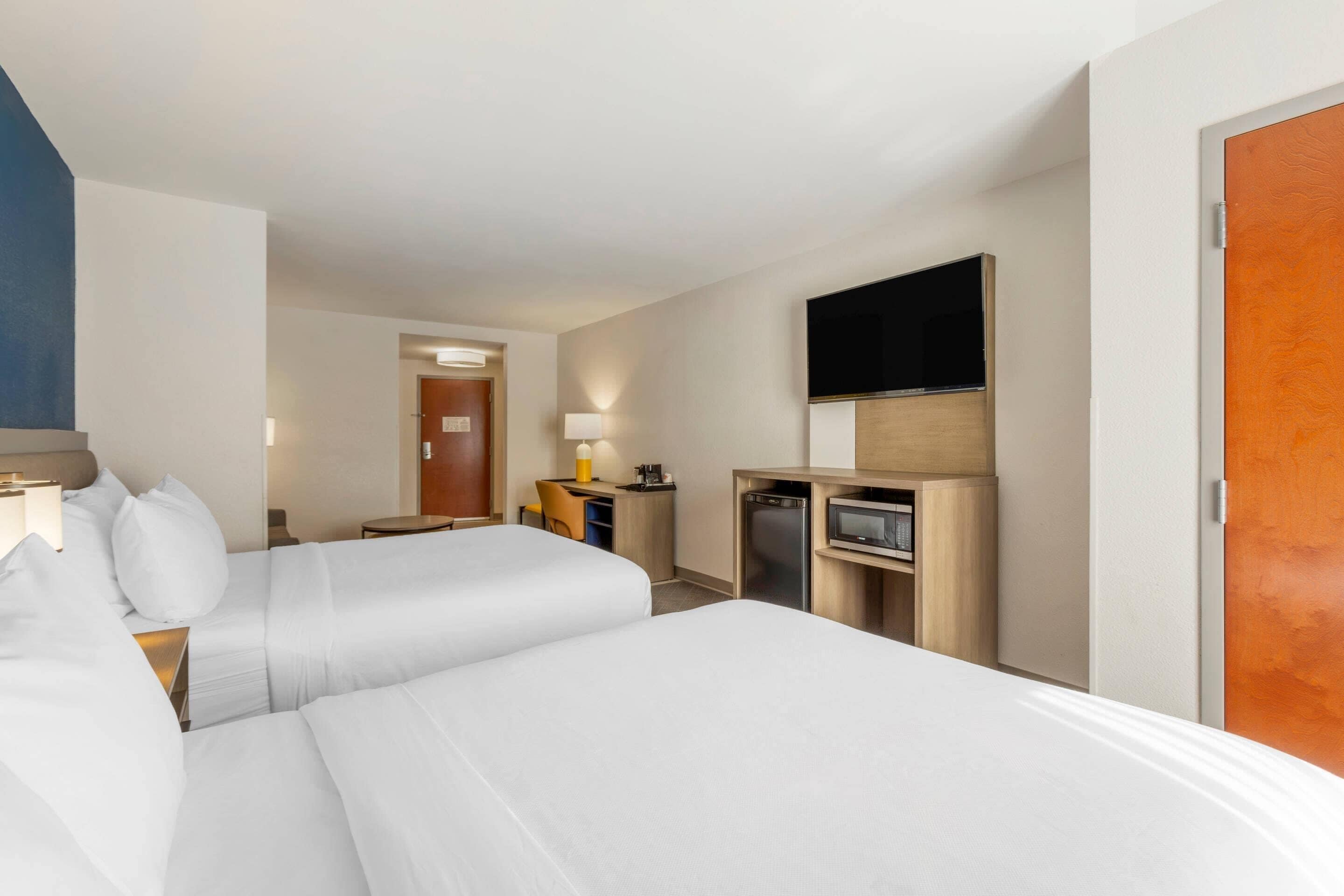 Una camera d'albergo con due letti, una scrivania con una lampada, un forno a microonde e una televisione a parete.