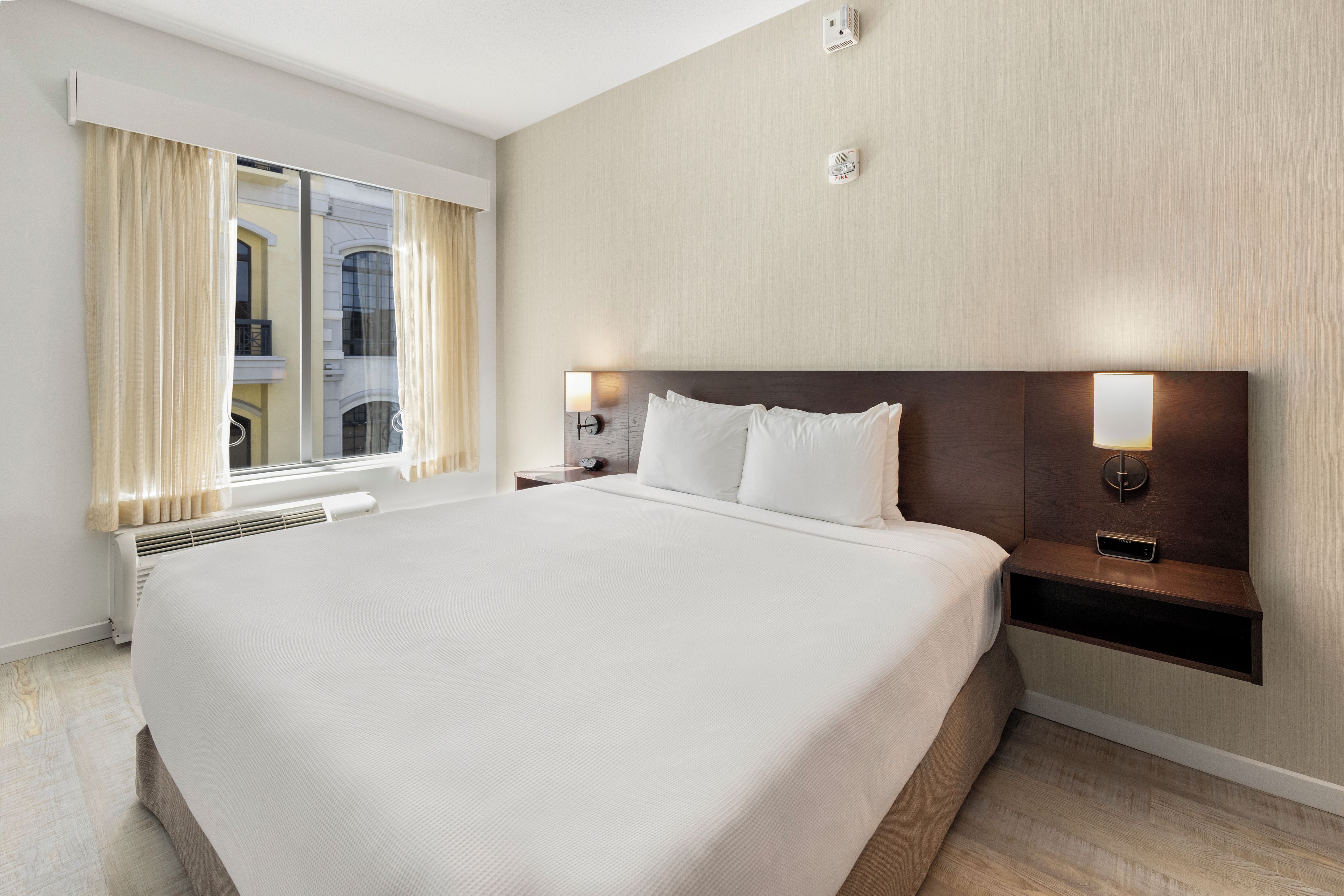 Une chambre d’hôtel avec un grand lit, une table de chevet avec une lampe, un réveil fixé au mur et une fenêtre avec des rideaux.