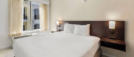 Une chambre d’hôtel avec un grand lit, une table de chevet avec une lampe, un réveil fixé au mur et une fenêtre avec des rideaux.