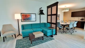 Una sala de estar con un sofá color turquesa, un sillón estampado, una mesa de centro y una lámpara de pie. En la pared hay un cuadro y un comedor con mesa y sillas.