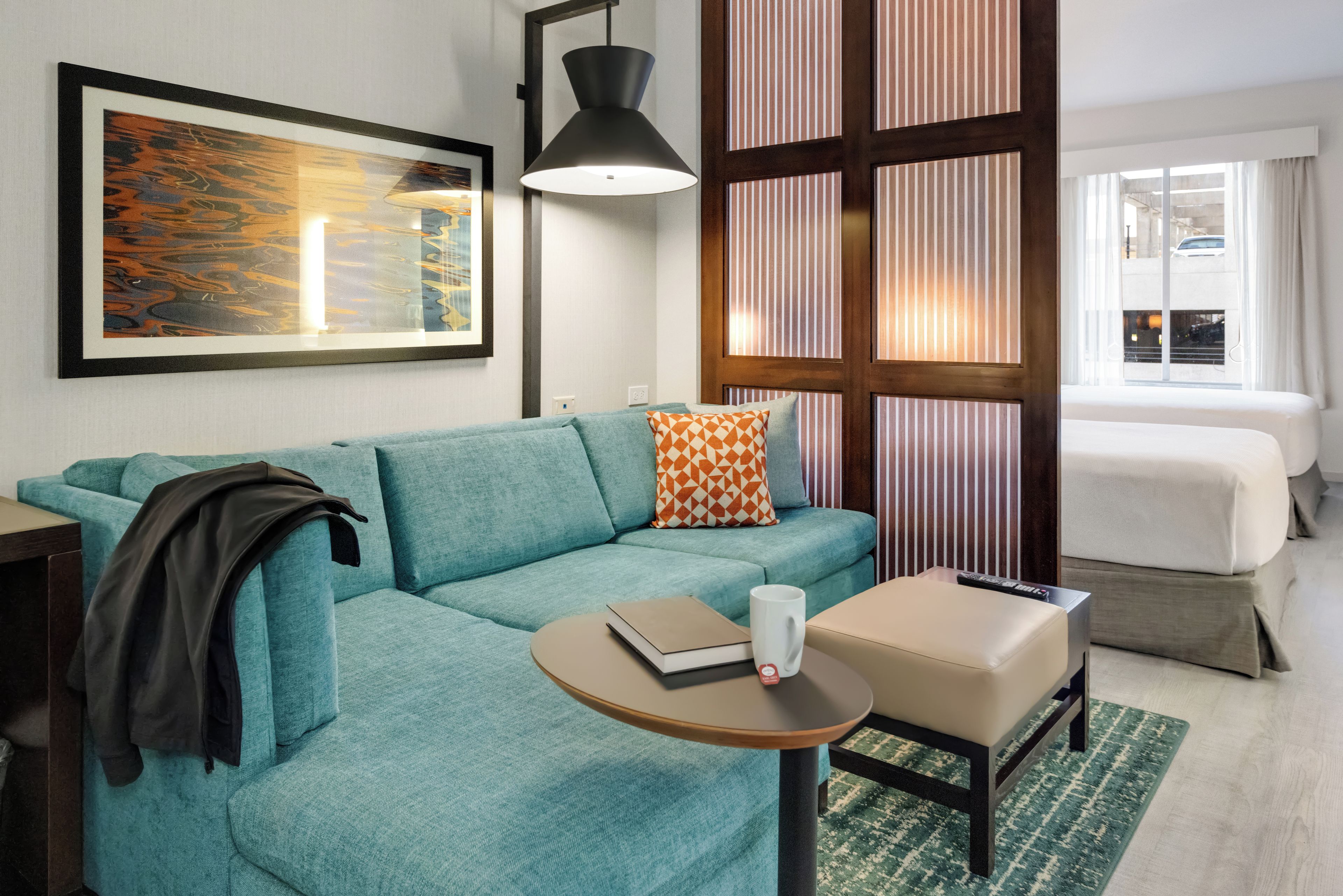 Una habitación de hotel moderna con un sofá color turquesa, una mesita redonda con un libro y una taza, una cama con cortinas a rayas y un cuadro abstracto grande en la pared.
