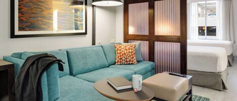 Ein modernes Hotelzimmer mit einem türkisfarbenen Sofa, einem kleinen runden Tisch mit Buch und Tasse, einem Bett mit gestreifteren Vorhängen und einem großen abstrakten Gemälde an der Wand.