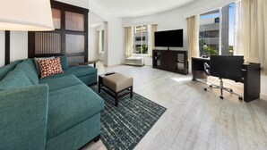 A modern living room with a sofa, ottoman, TV, and a desk with a chair. - Hyatt Place West Palm Beach/Downtown (West Palm Beach)