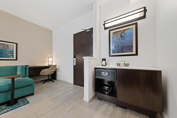 Habitación de hotel con un sofá verde azulado, una mesita, un mueble con cafetera, una obra de arte enmarcada y una puerta.