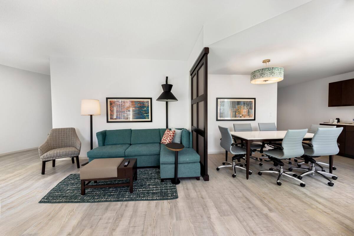 A modern living room with a teal sofa, a small side table, a floor lamp, a coffee table, and a dining area with a table and chairs.