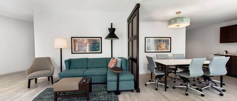 A modern living room with a teal sofa, a small side table, a floor lamp, a coffee table, and a dining area with a table and chairs.