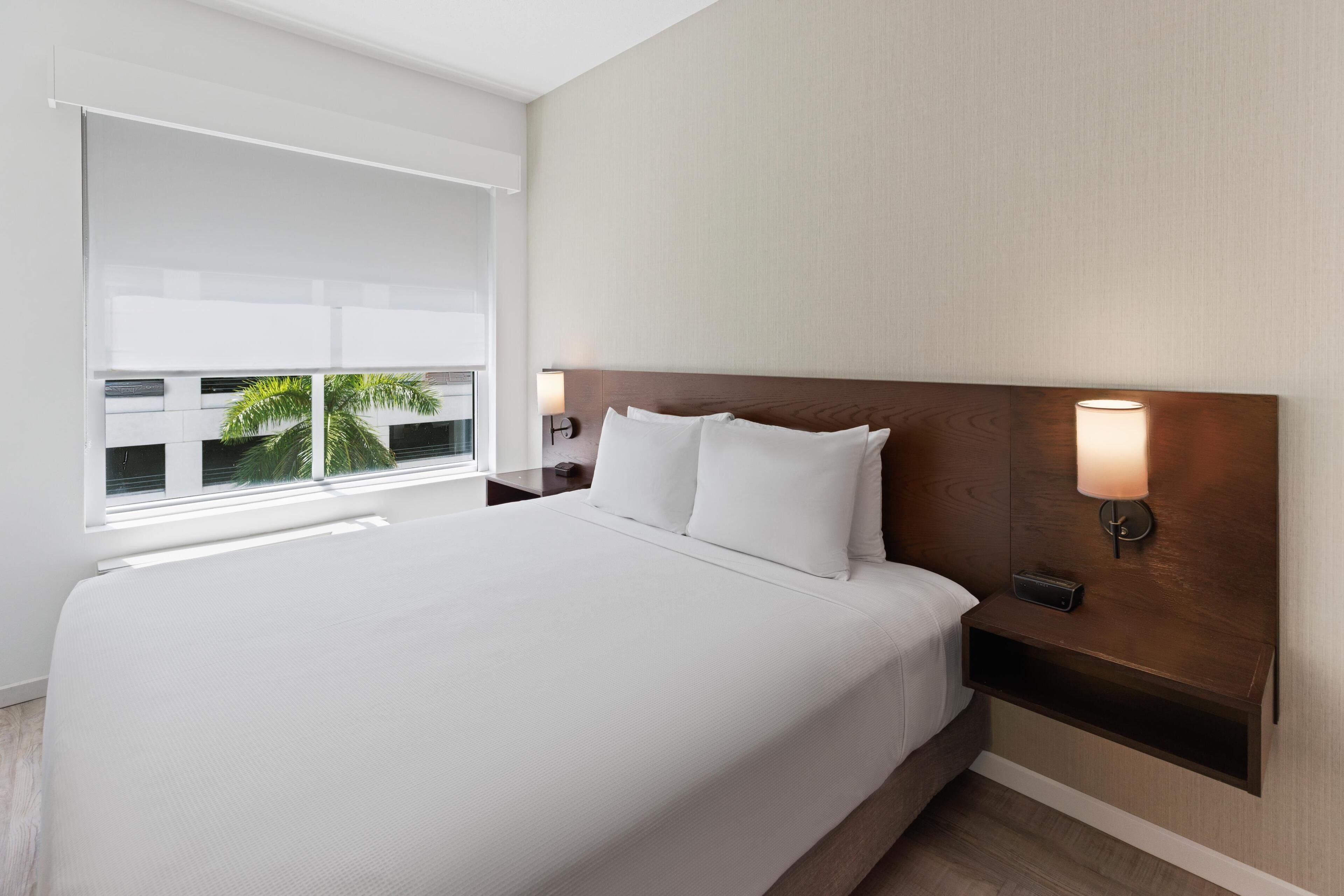 Une chambre d’hôtel avec un grand lit, une table de chevet avec une lampe, une fenêtre avec des stores et une vue sur des palmiers.