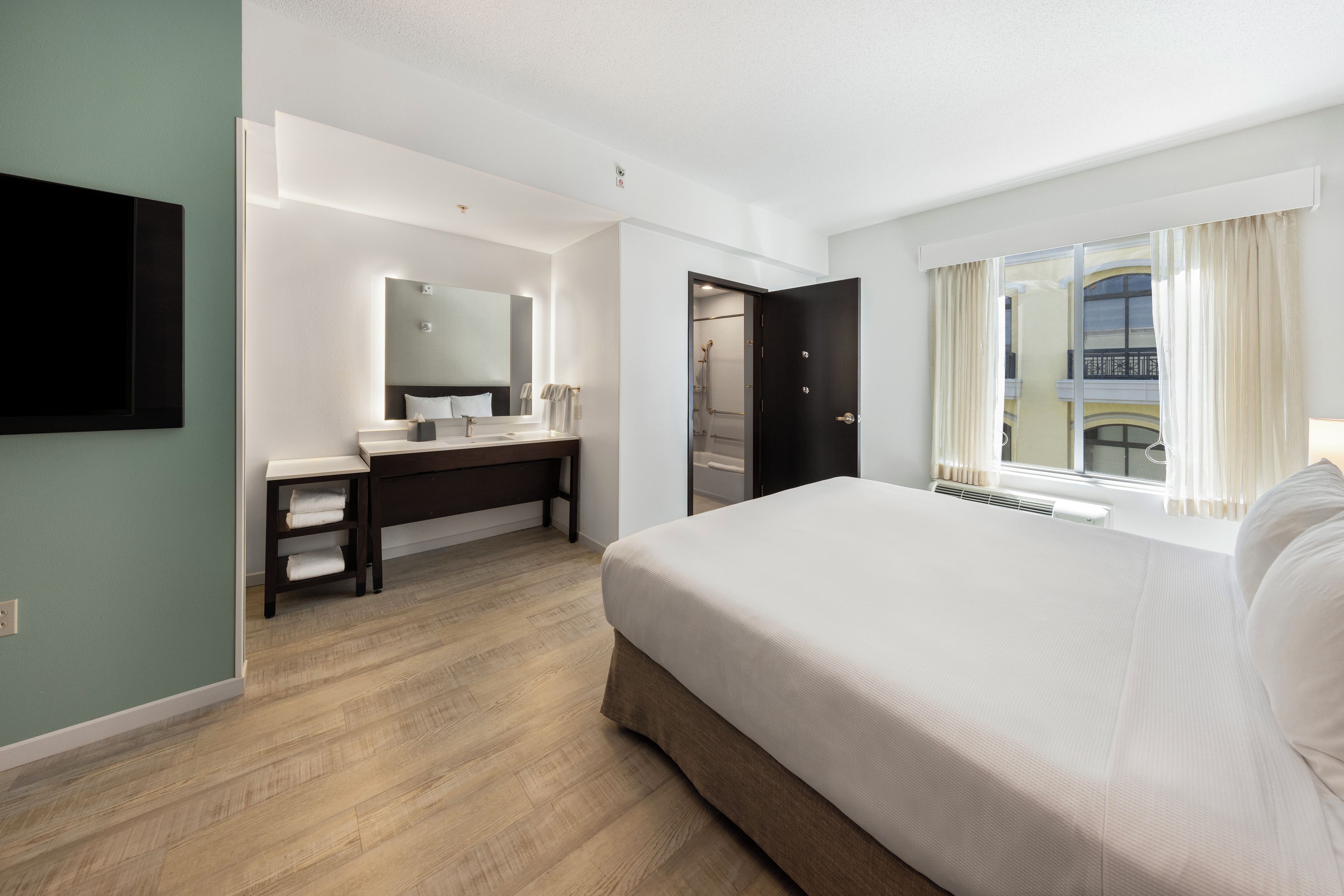 A hotel room with a large bed, a desk with a mirror, a flat-screen TV, and a window with curtains.