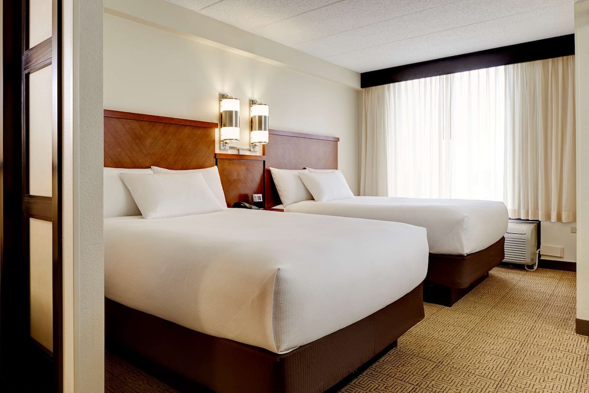 A hotel room with two beds, a wooden headboard, wall-mounted lights, and a large window with curtains.