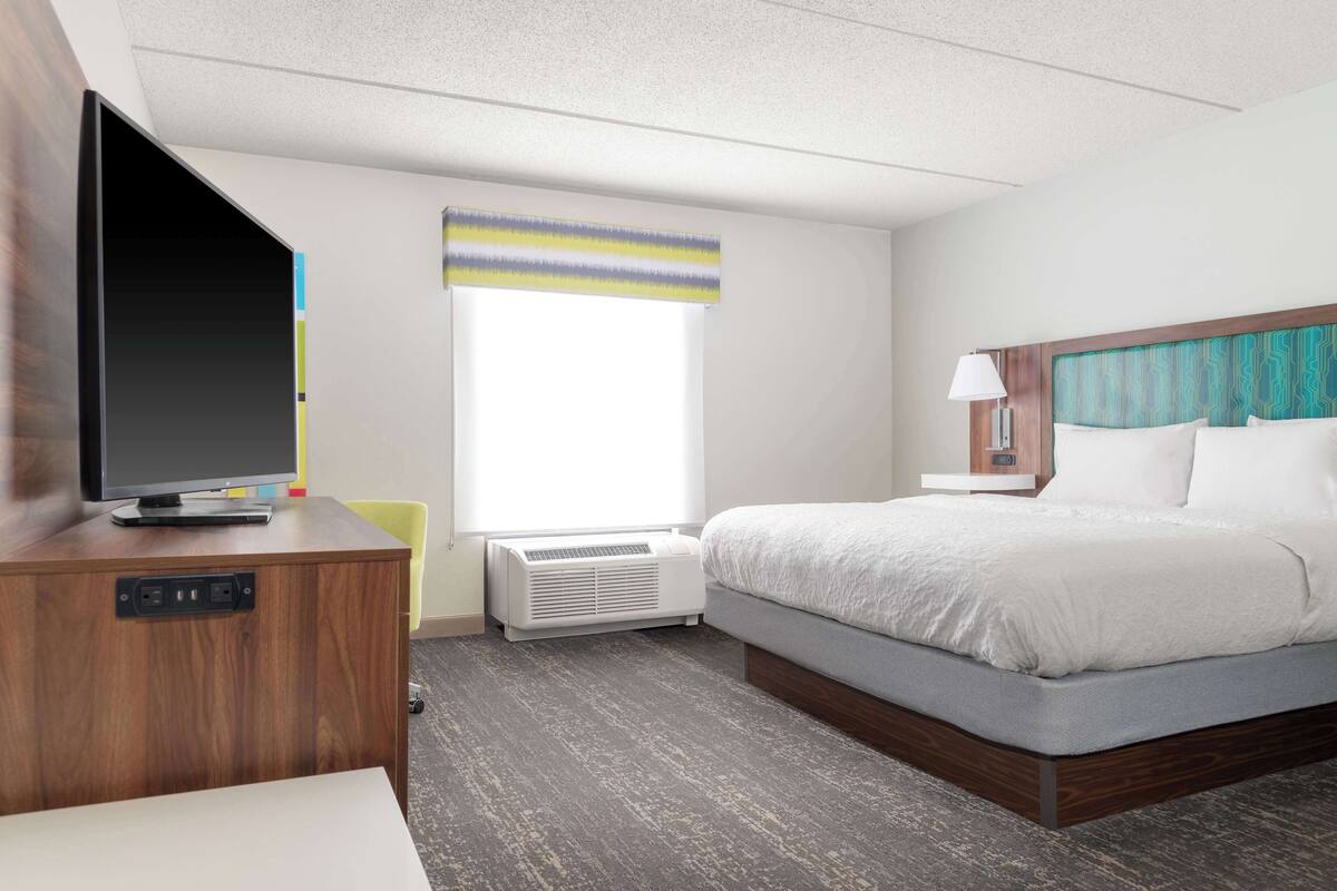 Quarto de hotel com cama, televisão sobre um armário de madeira, janela com cortina listrada e luminária fixada na parede.