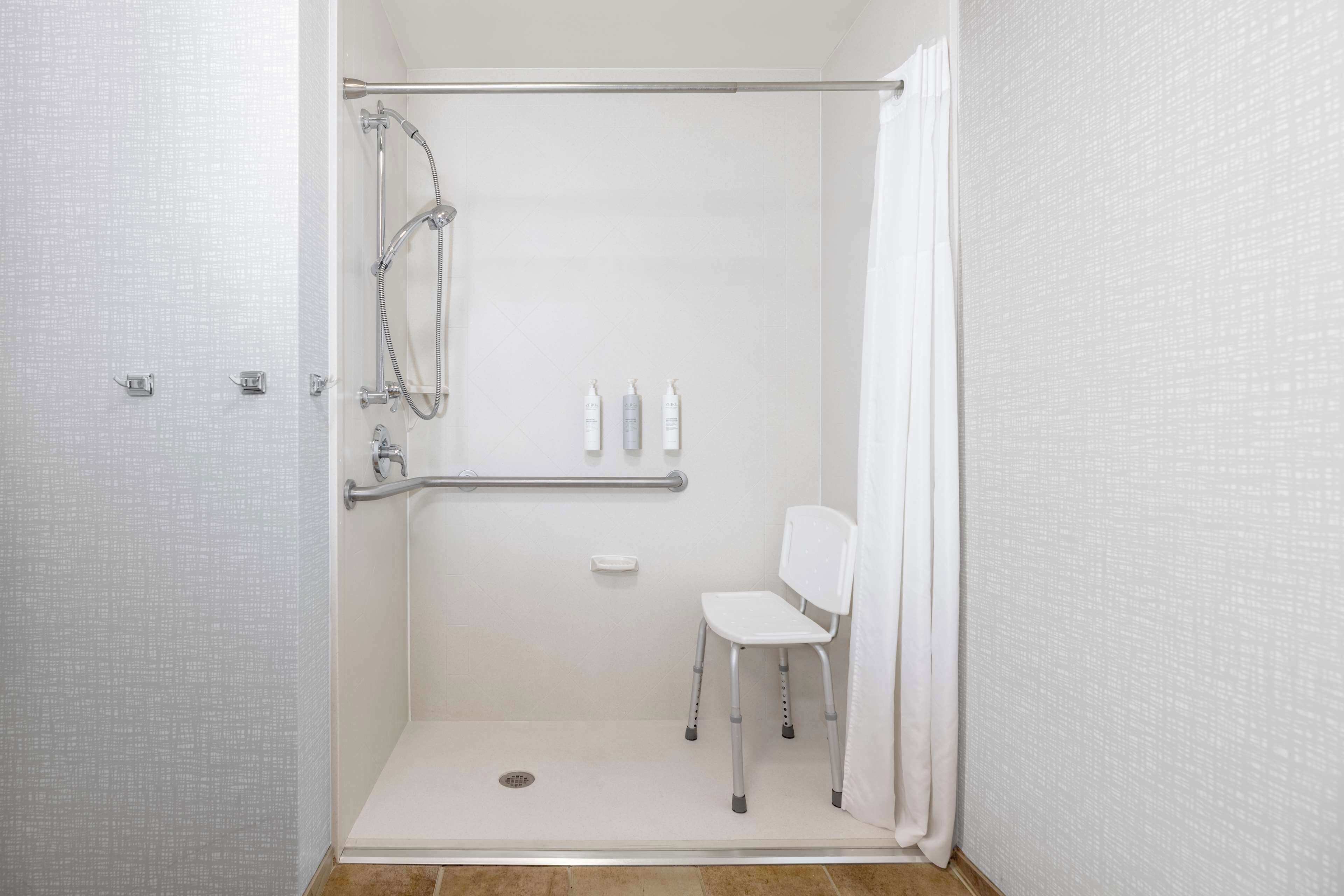 A wheelchair-accessible shower stall with a grab bar, a white chair, and a handheld showerhead.