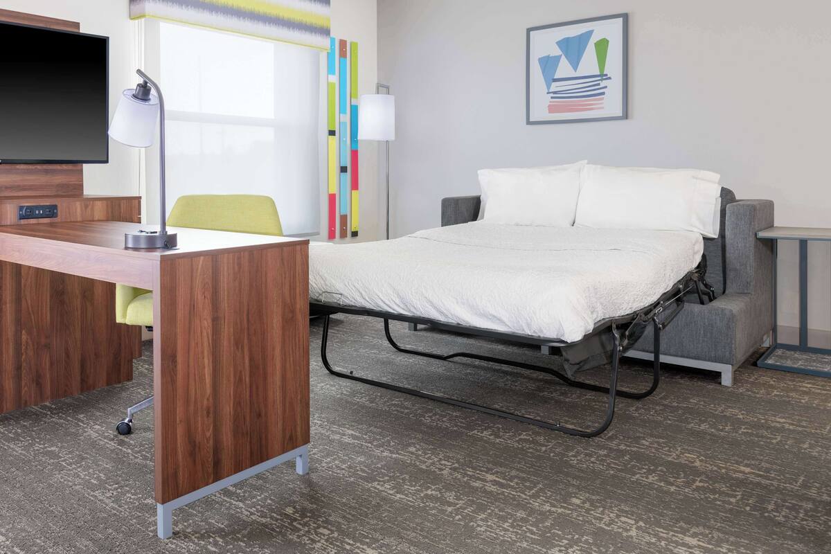 Quarto de hotel com cama, escrivaninha, cadeira e televisão.