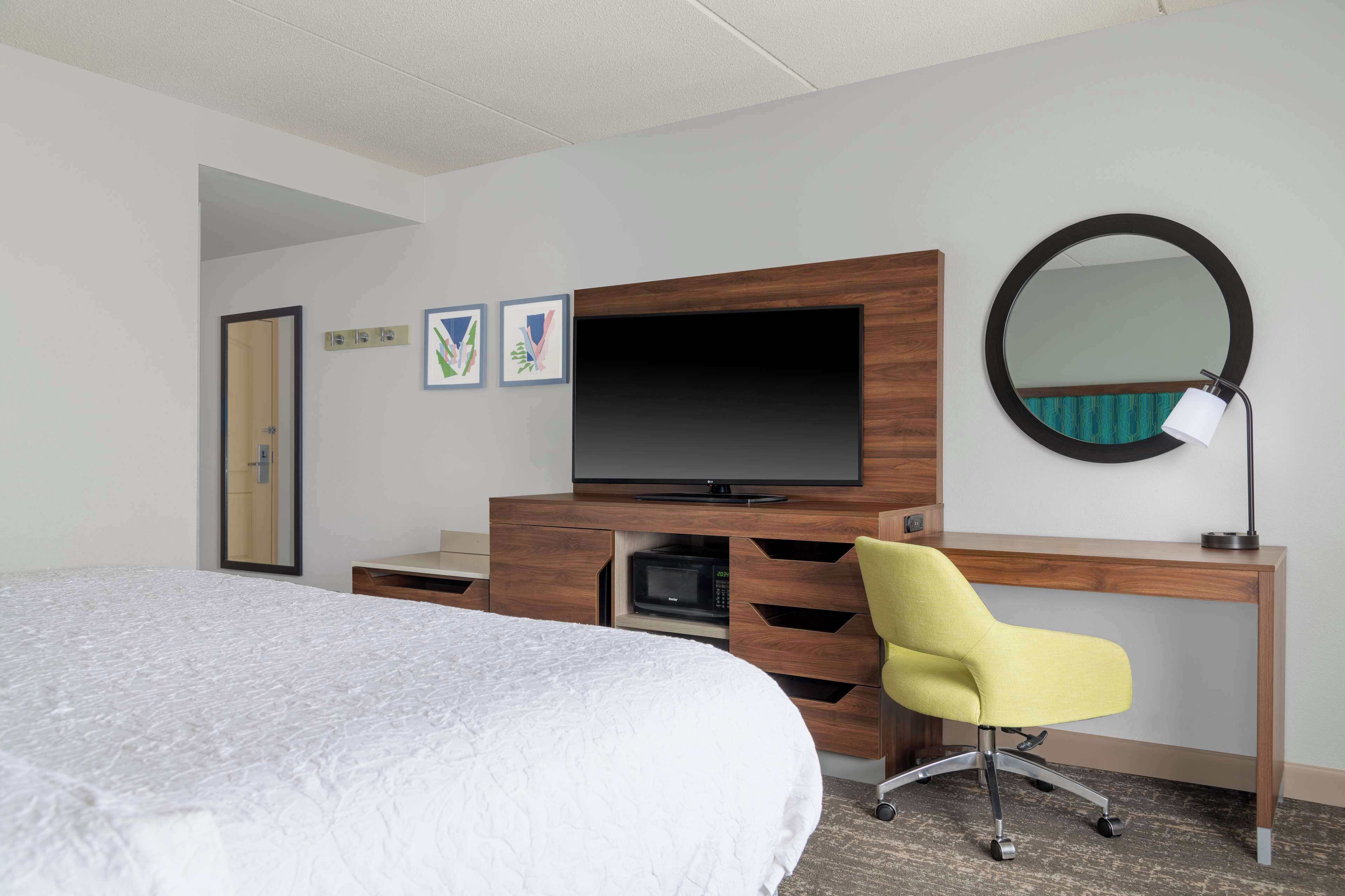 A hotel room with a bed, a wooden desk with a flat-screen TV, a yellow chair, a round mirror, and a microwave.