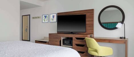 A hotel room with a bed, a wooden desk with a flat-screen TV, a yellow chair, a round mirror, and a microwave.