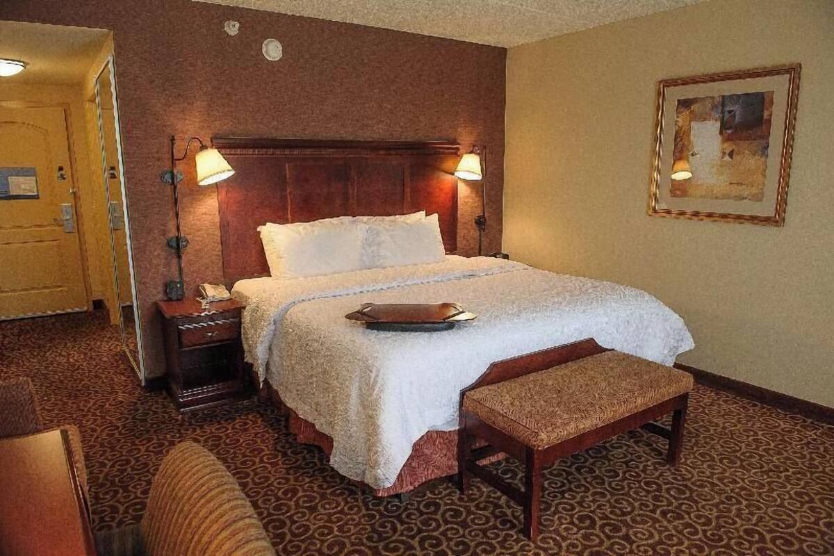 Quarto de hotel com uma cama grande, duas luminárias de cabeceira, um banco de madeira, uma mesinha de cabeceira e um quadro na parede.