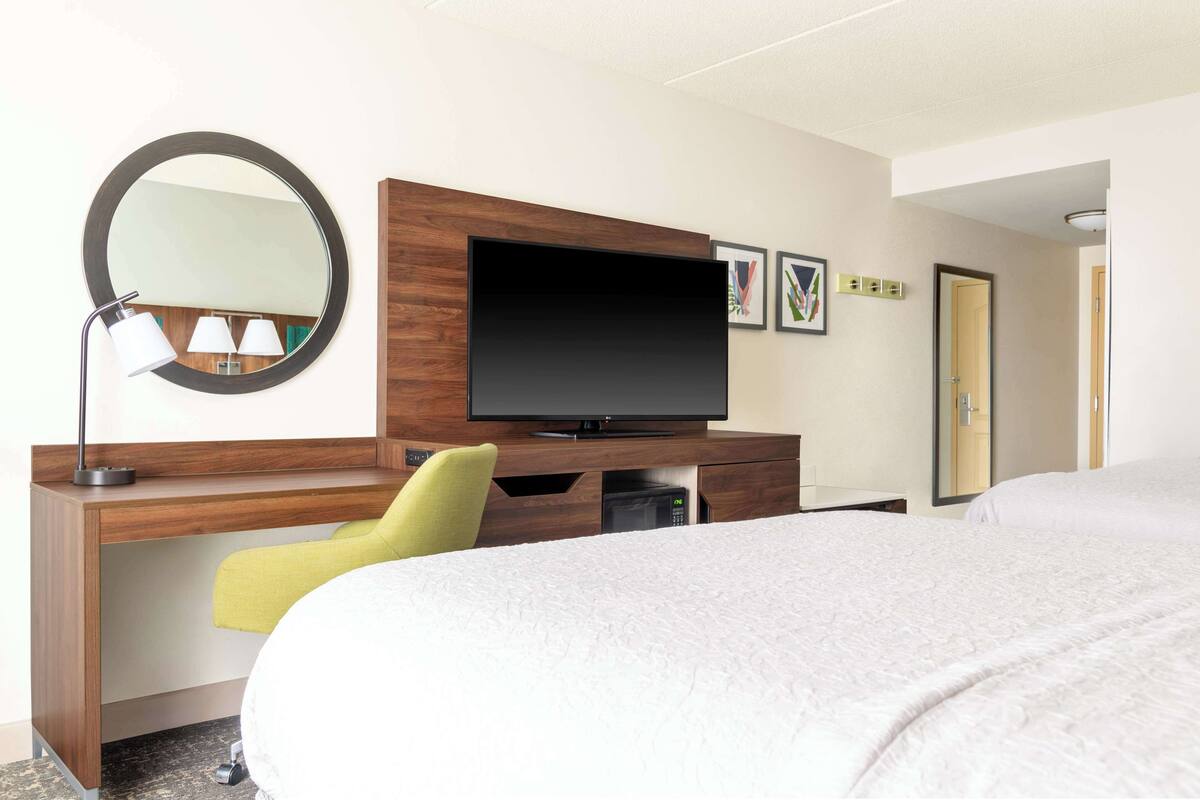 Quarto de hotel com uma cama, uma escrivaninha com televisão, um espelho e uma cadeira.