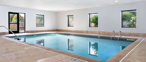 An indoor swimming pool with clear water, tiled floor, and windows offering views of greenery.